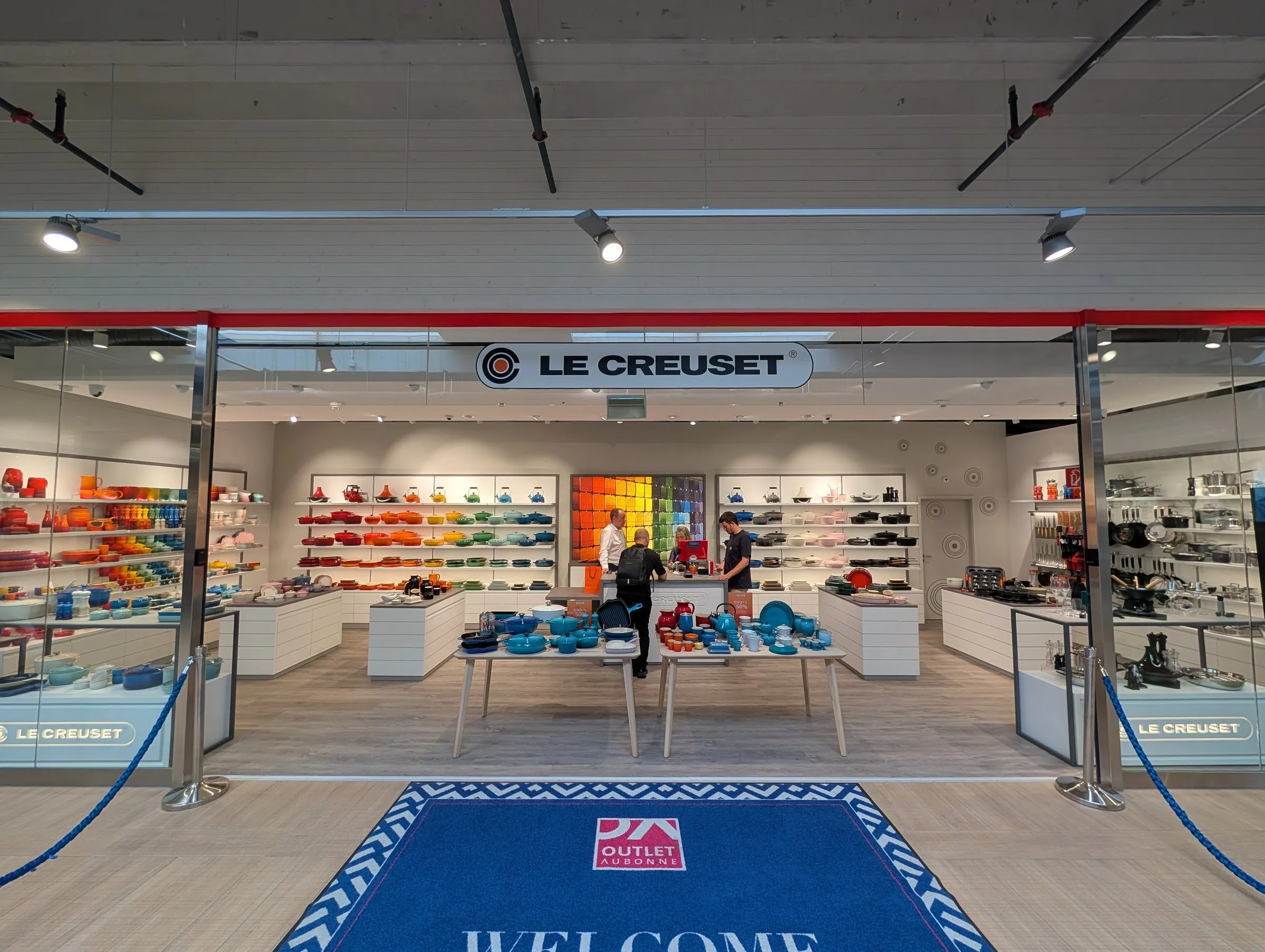 Devanture de la boutique Le Creuset à l’Aubonne Outlet, cadre de vitrine rouge et tapis d’accueil
