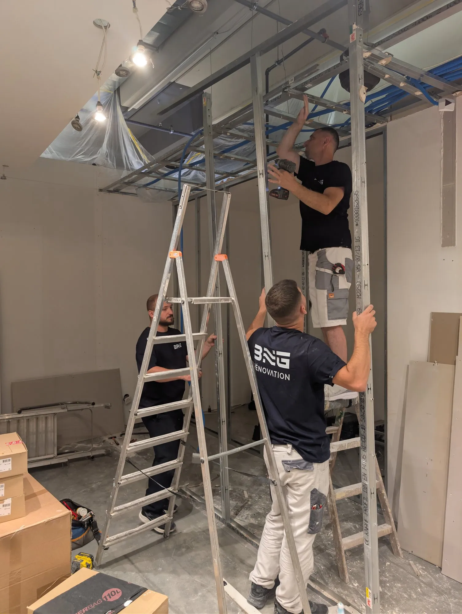 Deux ouvriers BNG en t-shirt au logo posent l’ossature métallique du faux-plafond